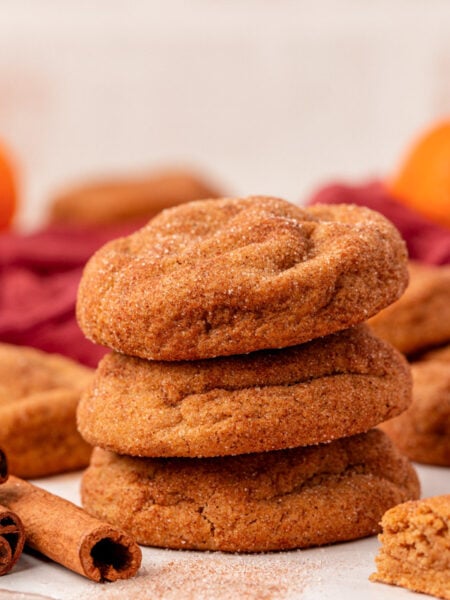 A stack of three pumpkin snickerdoodle cookies on a table with more cookies and cinnamon sticks around them.