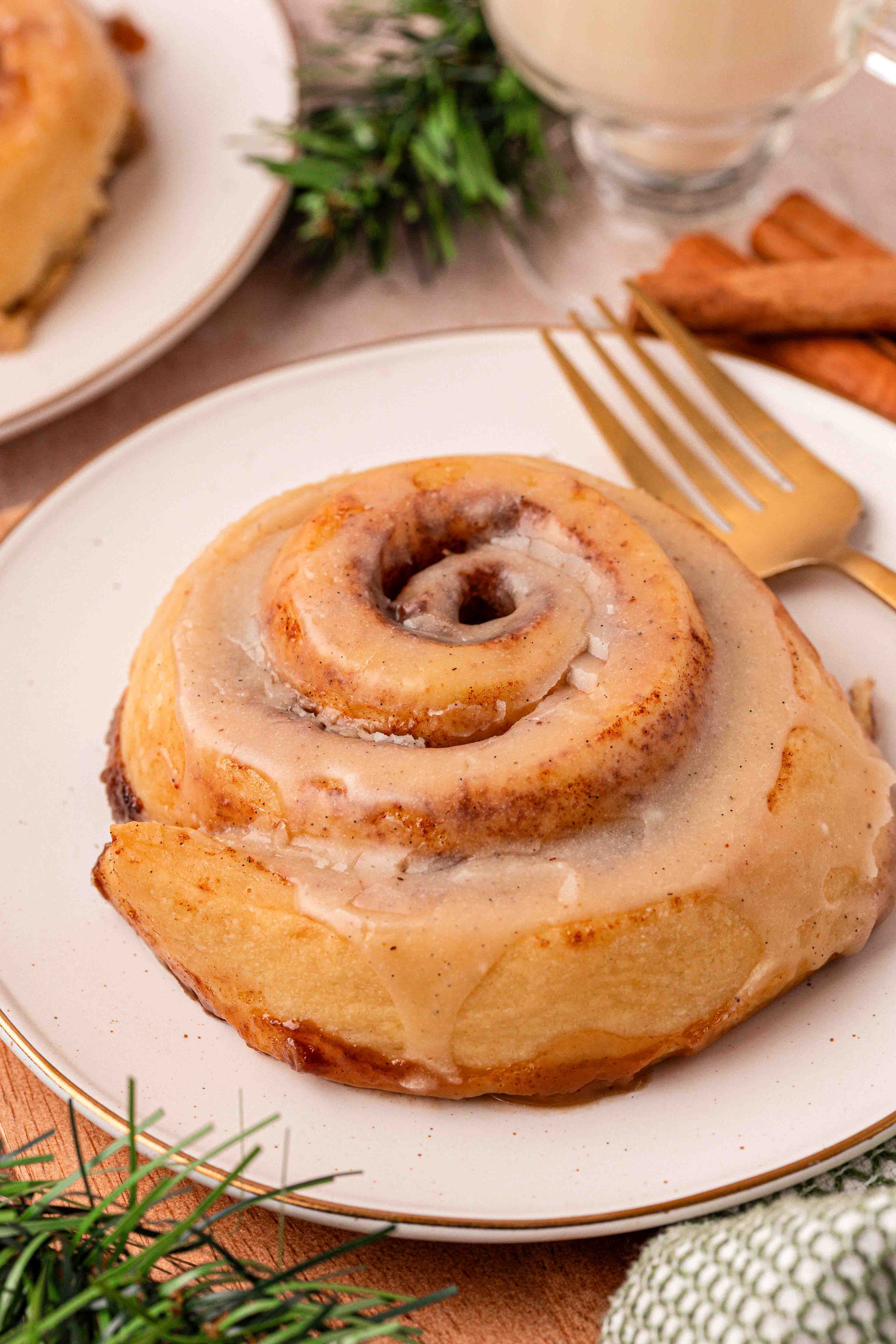 An eggnog cinnamon roll on a white plate with a gold fork next to it.