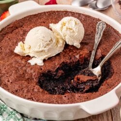 A dish of brownie pudding with two spoons in it a two scoops of vanilla ice cream on top.