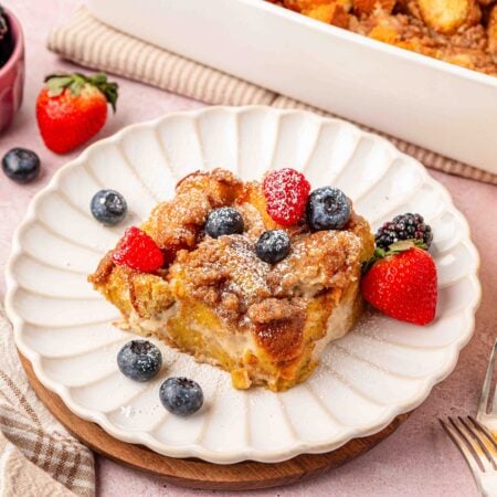 A slice of french toast casserole on a white scalloped plate with fresh berries on it.