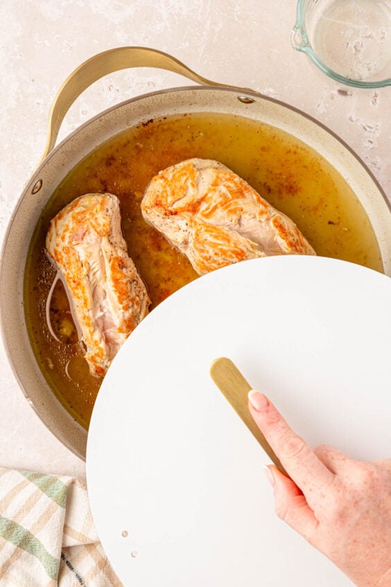 Chicken breasts and broth in a pan being covered with a lid.