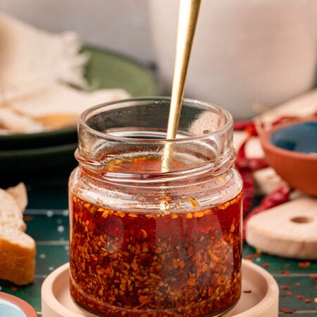 A jar of homemade chili onion crunch on a coaster with a spoon in it.