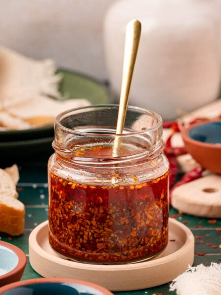 A jar of homemade chili onion crunch on a coaster with a spoon in it.