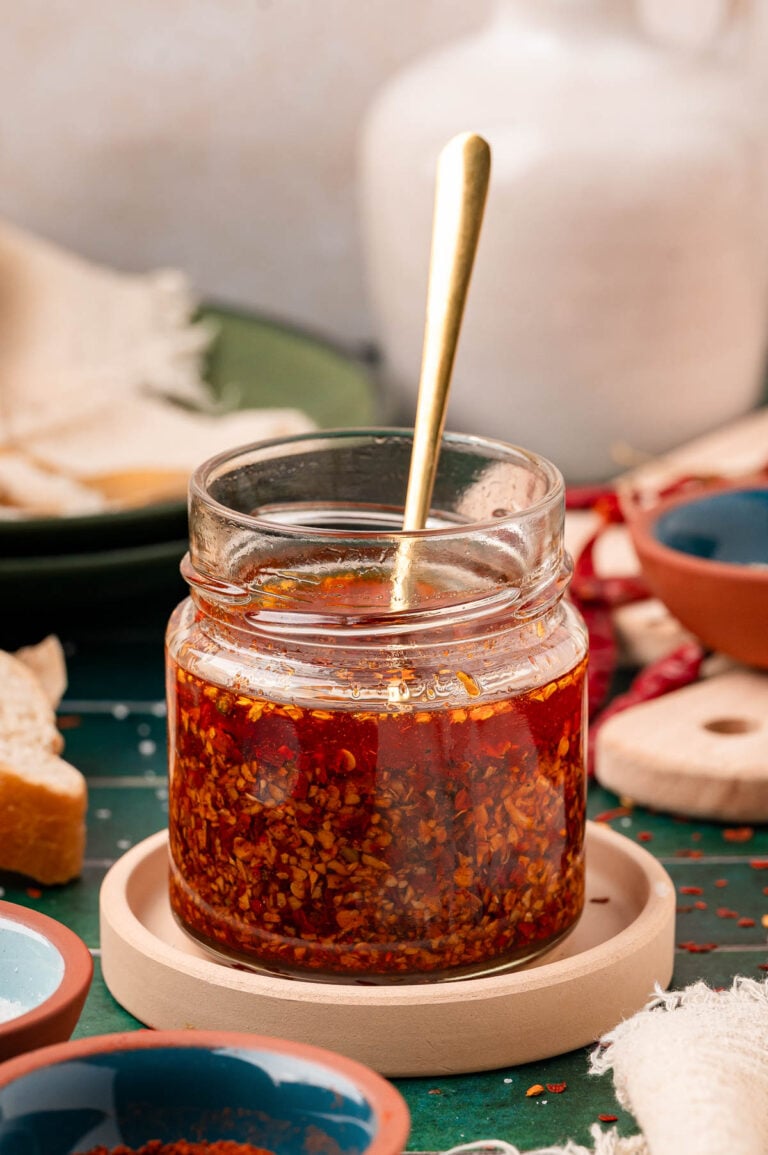 A jar of homemade chili onion crunch on a coaster with a spoon in it.