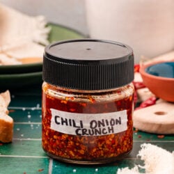 A jar of homemade chili onion crunch with a black top on a green table.