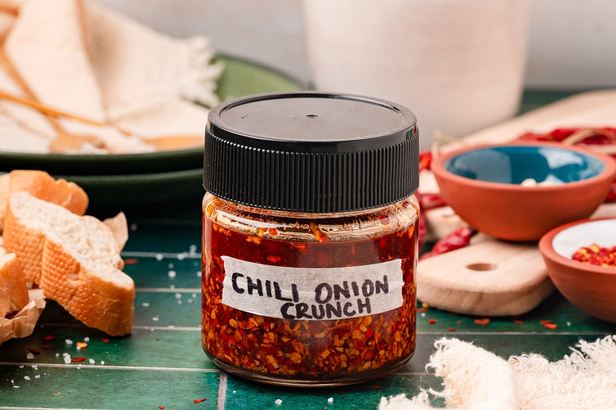 A jar of homemade chili onion crunch with a black top on a green table.