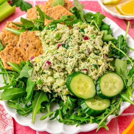 Chicken salad with green goddess dressing on a bed of arugula with crackers and cucumbers on a white plate.
