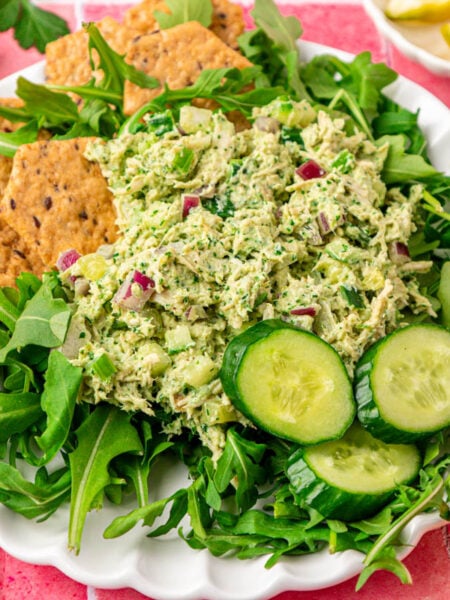 Chicken salad with green goddess dressing on a bed of arugula with crackers and cucumbers on a white plate.