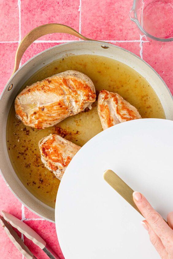 Chicken breast and stock in a pan with the cover being put on.