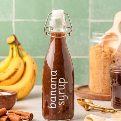 A bottle of banana coffee syrup on a table with bananas and brown sugar in the background.