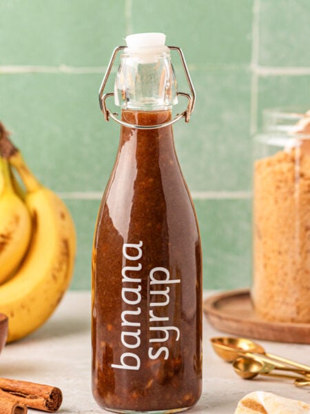 A bottle of banana coffee syrup on a table with bananas and brown sugar in the background.