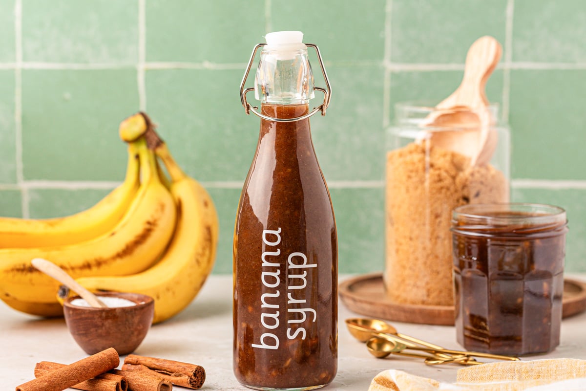 A bottle of banana coffee syrup on a table with bananas and brown sugar in the background.