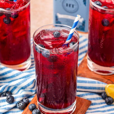 Three glasses of blueberry lemonade on a wood coaster on a blue and white striped cloth.