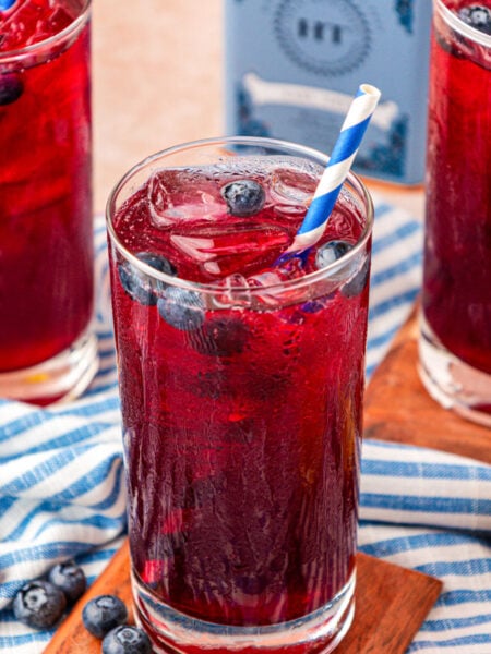 Three glasses of blueberry lemonade on a wood coaster on a blue and white striped cloth.