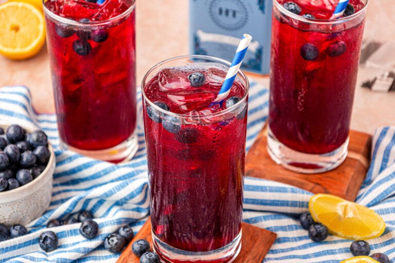 Three glasses of blueberry lemonade on a wood coaster on a blue and white striped cloth.